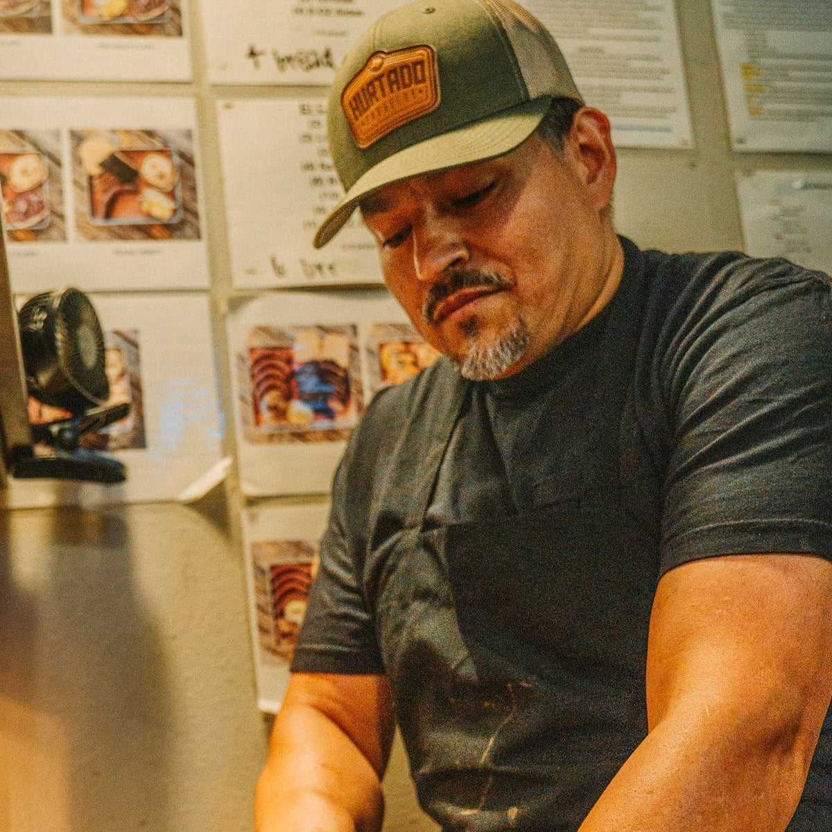 A Hurtado Barbecue pitmaster at the cutting board wearing the olive Leather Patch Trucker Hat